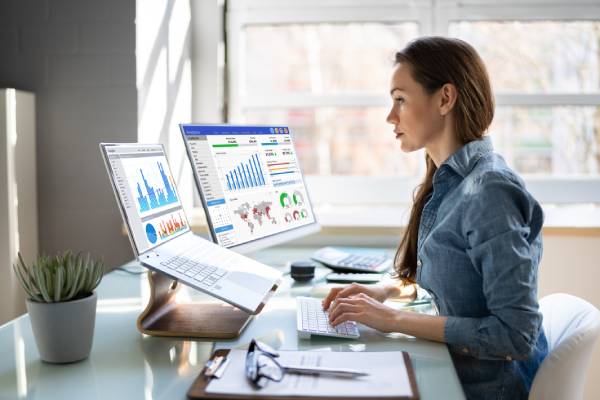 A Woman business analyst working in front of a desktop and laptop in an office room.